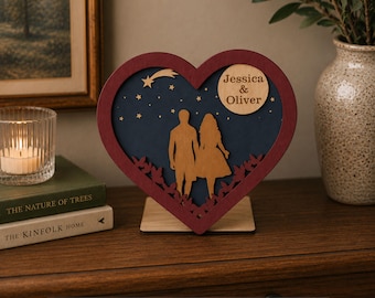 May include: A heart-shaped wooden decoration with a burgundy frame and a blue background. Inside, silhouettes of a couple holding hands are visible. The names "Jessica & Oliver" are engraved on a wooden circle. The text "Thickness: 1/8", 3mm, 4mm, 5mm" is at the bottom.