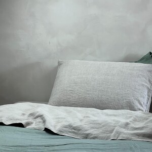 May include: A light gray linen pillow resting on a bed with a teal linen duvet cover.