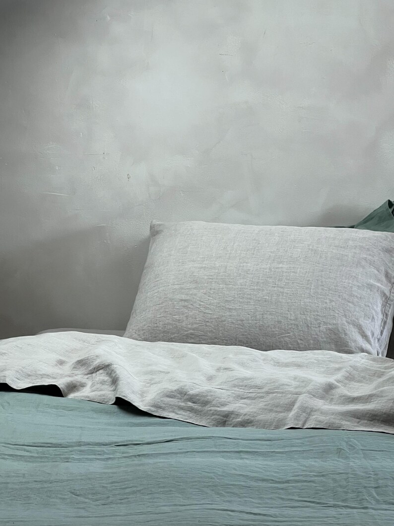 May include: A light gray linen pillow resting on a bed with a teal linen duvet cover.