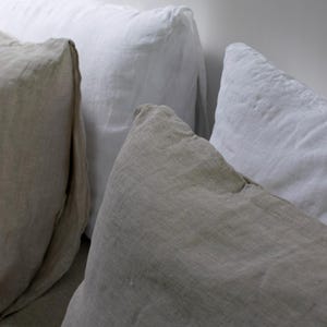 May include: Three pillows, one beige, one white, and one off-white, arranged on a bed. The pillows are made of linen and have a relaxed, casual look.