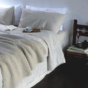 May include: A white linen bed with a gray knitted throw blanket, a book, and a cup of coffee on a wooden tray. The bed is against a white wall and there is a wooden chair with books on it in the corner.