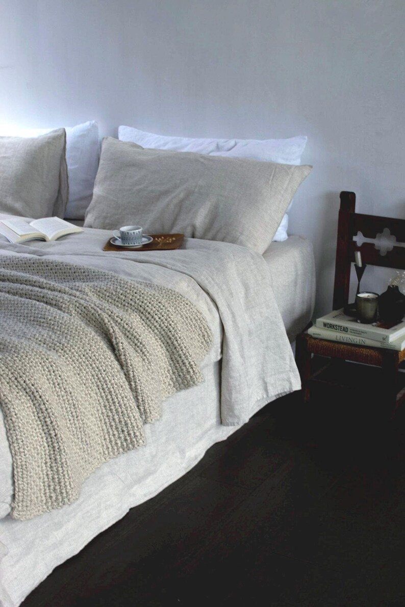 May include: A white linen bed with a gray knitted throw blanket, a book, and a cup of coffee on a wooden tray. The bed is against a white wall and there is a wooden chair with books on it in the corner.