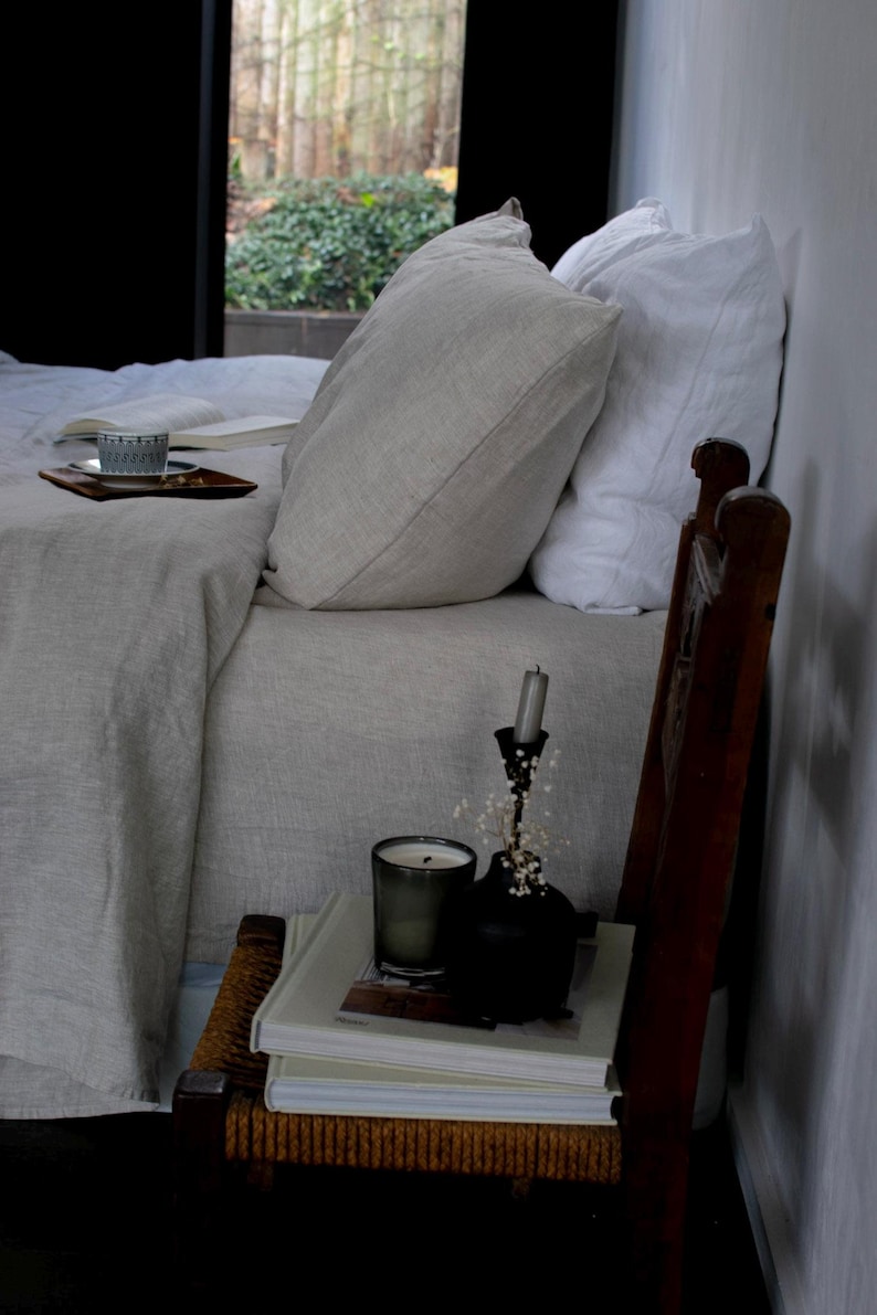 May include: A white linen bed with two pillows, a book, and a cup on a tray. A wooden chair with a stack of books and a candle holder with a lit candle sits next to the bed.