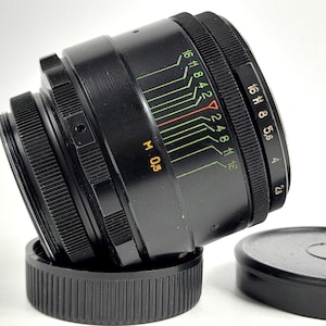May include: A black camera lens with focus and aperture settings. The lens has f-stop markings and a distance scale in metres. A black lens cap is next to the lens.