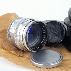May include: A vintage silver camera lens with black accents and a blue lens. The lens has markings and numbers, including "ЮПИТЕР-3" and "1:1.5 F=5cm". A black lens cap and a silver cap are also present.