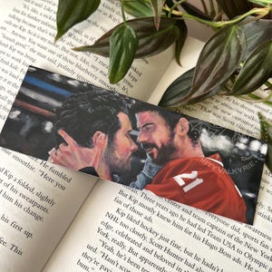 Könnte beinhalten: Lesezeichen mit einer Aquarellmalerei von zwei Männern in enger Umarmung. Einer der Männer trägt ein rotes Hockey-Trikot mit der Nummer 21. Das Lesezeichen liegt auf einem offenen Buch und grünem Laub.