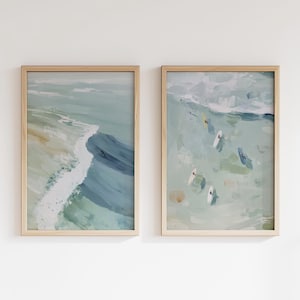 Puede incluir: Dos impresiones art&iacute;sticas abstractas enmarcadas. La impresi&oacute;n de la izquierda representa una ola rompiendo en la orilla, con tonos azules, verdes y blancos. La impresi&oacute;n de la derecha muestra surfistas en el oc&eacute;ano, con una paleta de colores similar. Ambas impresiones est&aacute;n en marcos de madera clara.