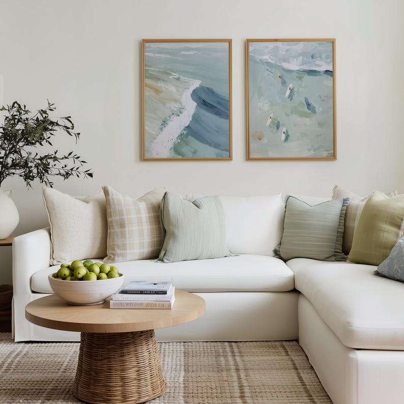 Puede incluir: Un sal&oacute;n con un sof&aacute; esquinero blanco y varios cojines en tonos neutros. Dos pinturas abstractas enmarcadas cuelgan de la pared. Una mesa de centro redonda de madera con base de mimbre sostiene un cuenco de manzanas verdes y libros.