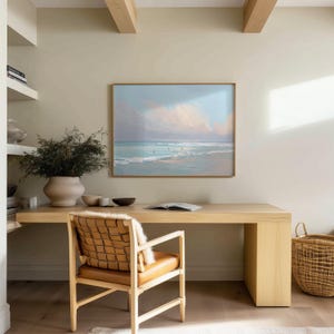 May include: A light wood desk with a framed beach scene painting, a woven leather chair, and a large beige vase with greenery. A woven basket sits on the floor. The room has a neutral color palette.