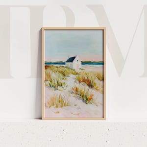 May include: Framed art print of a coastal scene. A white cottage with a pink door and dark blue roof sits on a sandy path, surrounded by tall grasses. The background features a blue ocean and a light blue and pink sky. The artwork is in a light wood frame.