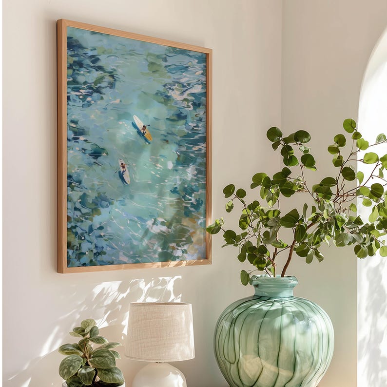 May include: A framed art print depicts two people paddleboarding on turquoise water. The artwork is in a light wood frame and hangs on a white wall. A green vase with leafy branches and a small lamp are in the foreground.