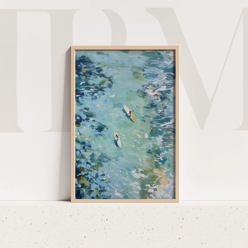 May include: A framed art print featuring an aerial view of two people paddleboarding on turquoise water. The artwork has a light wooden frame and depicts a serene scene with blue and green hues, evoking a sense of calm and leisure.