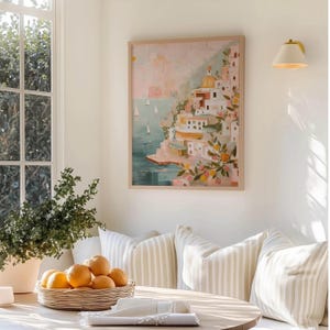 K&ouml;nnte beinhalten: Gerahmtes Gem&auml;lde eines italienischen K&uuml;stendorfes mit wei&szlig;en Geb&auml;uden, blauem Meer und Segelbooten. Das Kunstwerk befindet sich in einem hellen Holzrahmen und h&auml;ngt an einer wei&szlig;en Wand. Ein Korb mit Orangen steht auf einem Holztisch vor gestreiften Kissen.