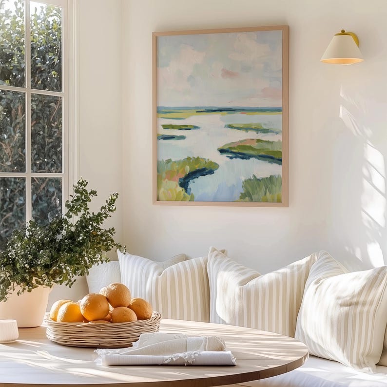 May include: A framed landscape painting with a light wood frame hangs on a white wall. The painting depicts a marsh scene with blue water, green and yellow land masses, and a light blue and pink sky. A basket of oranges sits on a round wooden table.