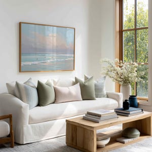 May include: A living room scene featuring a white sofa adorned with various throw pillows in shades of green, blue, and pink. A framed landscape painting hangs above the sofa. A wooden coffee table holds books and decorative items. A large window provides natural light.