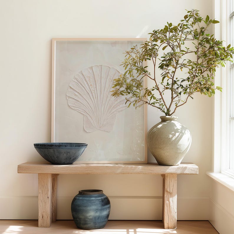 K&ouml;nnte beinhalten: Eine ruhige Innenszene mit einer hellen Holzbank, auf der sich eine blaue Sch&uuml;ssel und eine kleine blaue Vase befinden. Ein gro&szlig;es gerahmtes Kunstwerk mit einem wei&szlig;en Muscheldesign h&auml;ngt dar&uuml;ber. Eine runde, hellgr&uuml;ne Vase enth&auml;lt belaubte Zweige.