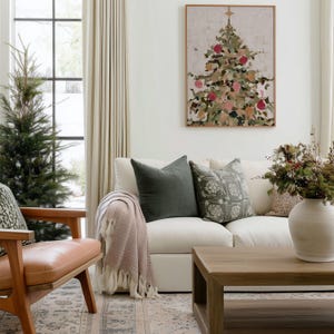 May include: A living room scene featuring a white sofa with green and patterned pillows, a wooden coffee table, and a framed Christmas tree painting. A pink throw blanket with fringe is draped over the sofa. A wooden chair with a leather seat is on the left.