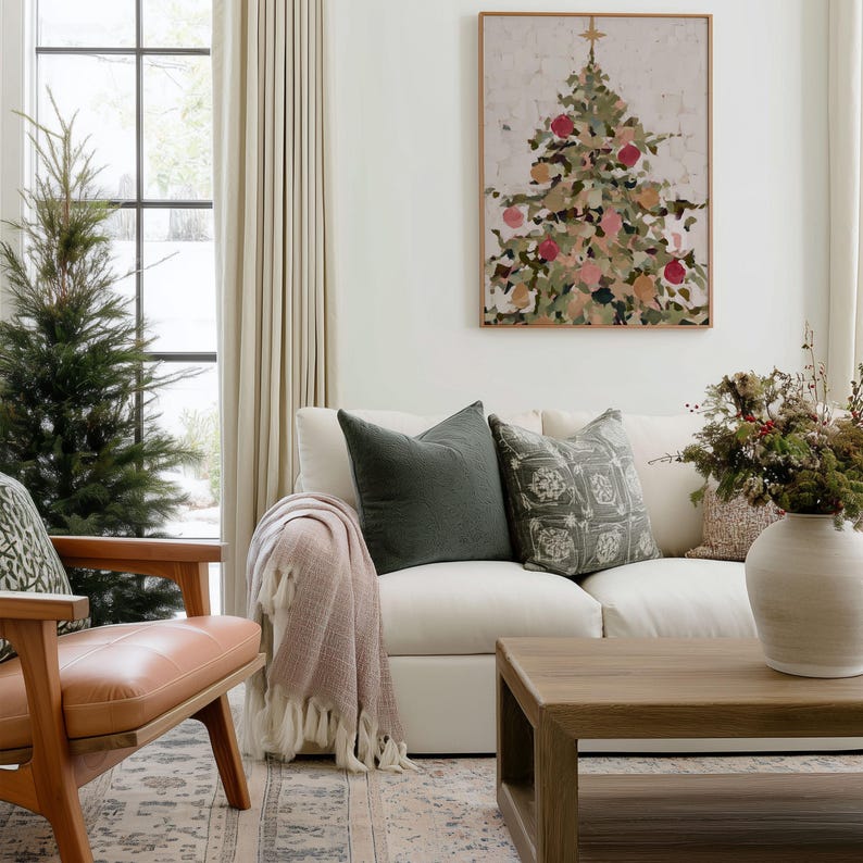 May include: A living room scene featuring a white sofa with green and patterned pillows, a wooden coffee table, and a framed Christmas tree painting. A pink throw blanket with fringe is draped over the sofa. A wooden chair with a leather seat is on the left.