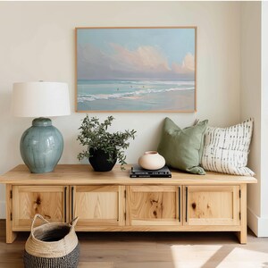 May include: A light wood cabinet with four doors, topped with decor including a blue-green lamp, a black vase with greenery, a small pink vase, and two pillows. A framed painting of a beach scene hangs above the cabinet. A woven basket sits on the floor.