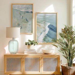 Puede incluir: Decoraci&oacute;n de sal&oacute;n de tem&aacute;tica costera con dos impresiones art&iacute;sticas abstractas enmarcadas con escenas oce&aacute;nicas en tonos azules y verdes. Una l&aacute;mpara de cristal azul claro con una pantalla beige se encuentra sobre un mueble de madera clara con paneles de puerta tejidos. Una planta en maceta y un cuenco blanco completan la escena.