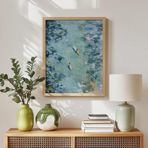 May include: A framed art print depicts two people paddleboarding on a turquoise body of water. The artwork is in a light wood frame and hangs above a wooden cabinet. A green vase, a white and green vase, a lamp, and books are on the cabinet.