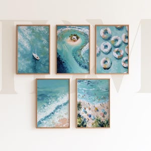 May include: Five framed art prints with a coastal theme. The paintings depict a sailboat, an island, pool floats, ocean waves, and a beach scene. The artwork features shades of blue, green, and tan, with wooden frames.