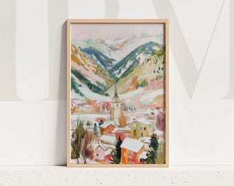 May include: A framed watercolour painting depicting a snow-covered village in a valley between mountains. The artwork shows a church spire, colourful buildings, and snow-capped peaks. The frame is a light wood colour.