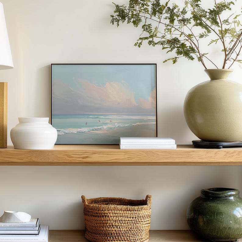 May include: A coastal-themed display featuring a framed seascape painting with a black frame, a white vase, a large olive-green vase, and a woven basket. The painting depicts a beach scene with soft pastel colors.