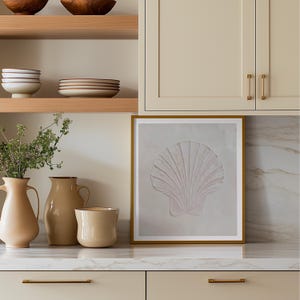 K&ouml;nnte beinhalten: Ein gerahmtes Kunstwerk mit einem strukturierten, hellrosa Muscheldesign. Das Kunstwerk wird auf einer Arbeitsplatte mit neutralen K&uuml;chenelementen ausgestellt, darunter Vasen, Geschirr und Schr&auml;nke mit goldenen Griffen. Die Gesamt&auml;sthetik ist k&uuml;sten- und minimalistisch.