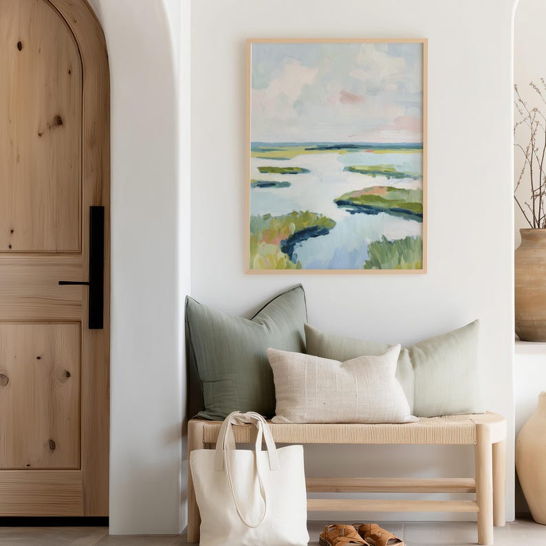 May include: An abstract landscape painting in a light wood frame hangs above a woven bench with sage green and cream pillows. A white tote bag and brown leather sandals sit below the bench, creating a serene entryway scene.