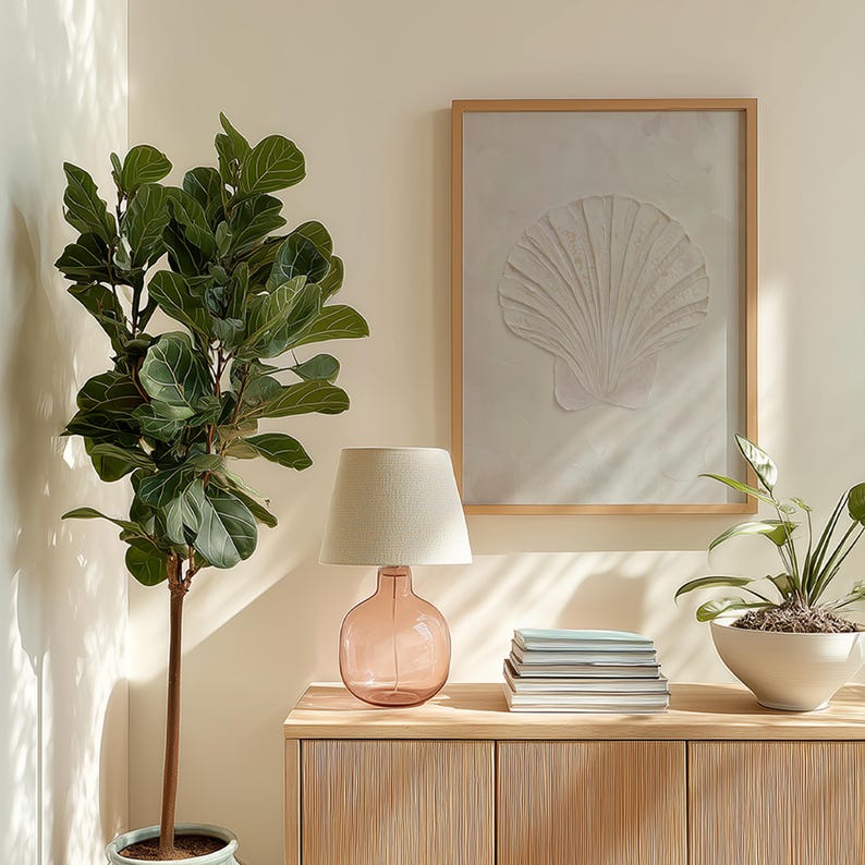 K&ouml;nnte beinhalten: Eine Innenraumszene mit K&uuml;sten-Thema, mit einer rosa Glas-Tischlampe mit beigem Lampenschirm, einem gerahmten Muschel-Kunstwerk und einer gro&szlig;en Topfpflanze. Die Lampe steht auf einem hellen Holzschrank, mit B&uuml;chern und einer kleinen Topfpflanze.
