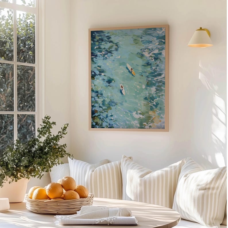 May include: A framed art print depicts two people paddleboarding on turquoise water. The artwork is in a light wood frame and hangs on a white wall. The room also features a window, a basket of oranges, and striped pillows.