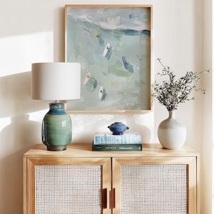 Puede incluir: Un mueble de madera clara con puertas tejidas sostiene una l&aacute;mpara de cer&aacute;mica verde y azul con una pantalla blanca. Una pintura enmarcada de surfistas en el oc&eacute;ano cuelga sobre el mueble. Un jarr&oacute;n blanco con vegetaci&oacute;n y un cuenco decorativo azul est&aacute;n encima.