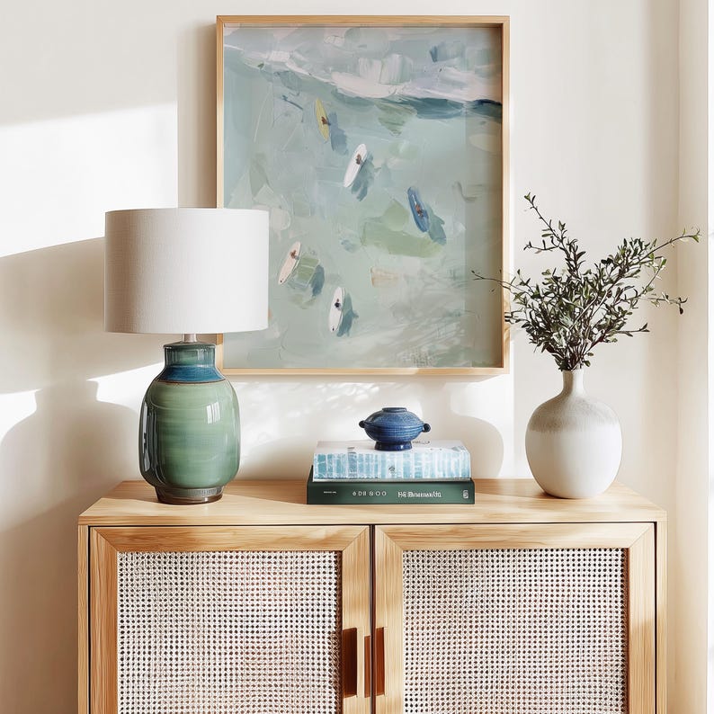 Puede incluir: Un mueble de madera clara con puertas tejidas sostiene una l&aacute;mpara de cer&aacute;mica verde y azul con una pantalla blanca. Una pintura enmarcada de surfistas en el oc&eacute;ano cuelga sobre el mueble. Un jarr&oacute;n blanco con vegetaci&oacute;n y un cuenco decorativo azul est&aacute;n encima.