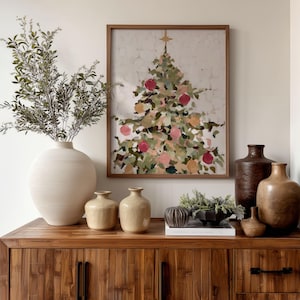 May include: A wooden cabinet displays a framed Christmas tree painting with red and pink ornaments. A large white vase holds leafy branches. Several ceramic vases and a decorative bowl with greenery complete the holiday decor.