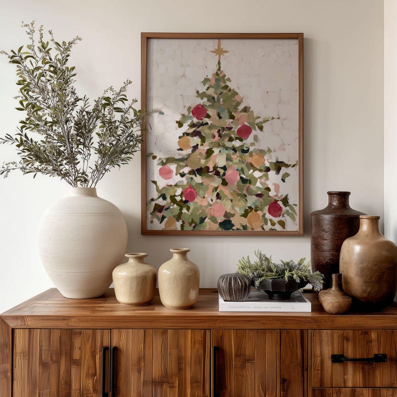 May include: A wooden cabinet displays a framed Christmas tree painting with red and pink ornaments. A large white vase holds leafy branches. Several ceramic vases and a decorative bowl with greenery complete the holiday decor.