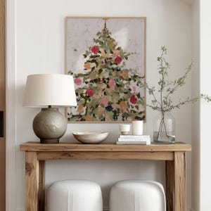 May include: A framed abstract painting of a Christmas tree with red, pink, and gold ornaments hangs above a wooden console table. A gray lamp with a white shade, a decorative bowl, and candles are on the table. Two white upholstered stools are below.