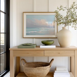 May include: A light wood console table with a framed beach scene painting, a green bowl, and a large beige vase with greenery. A woven basket sits below the drawers. The room has white paneling and a black window frame.
