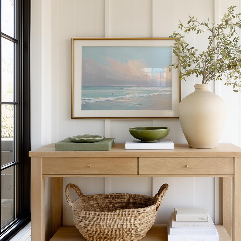 May include: A light wood console table with a framed beach scene painting, a green bowl, and a large beige vase with greenery. A woven basket sits below the drawers. The room has white paneling and a black window frame.