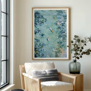 May include: A framed art print depicts a serene waterscape with two figures on surfboards. The artwork features a palette of blues and greens, with a light brown wooden frame. The print hangs above a white armchair with a striped pillow.