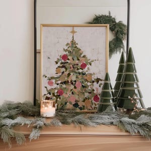May include: A framed Christmas tree painting with red and pink ornaments is displayed on a wooden surface. Beside it are three green ceramic Christmas tree decorations. A lit candle and garland complete the festive scene.