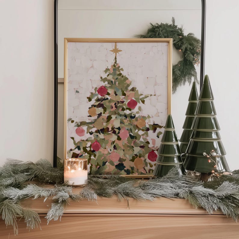 May include: A framed Christmas tree painting with red and pink ornaments is displayed on a wooden surface. Beside it are three green ceramic Christmas tree decorations. A lit candle and garland complete the festive scene.