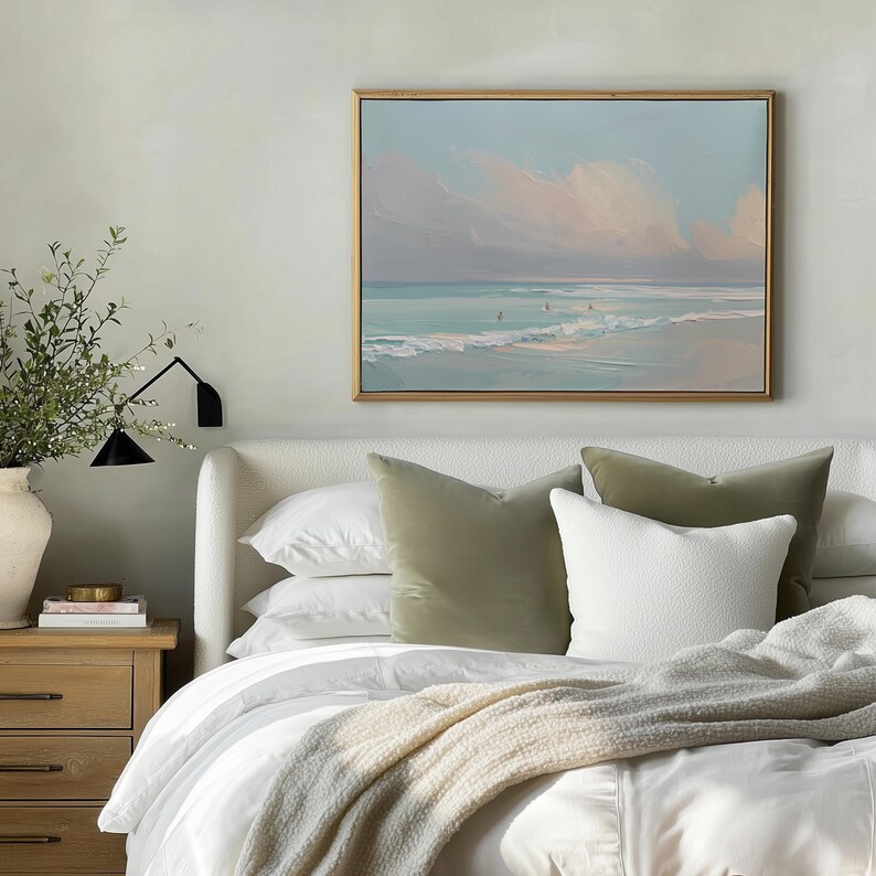May include: A bedroom scene featuring a bed with white linens, olive green and white pillows, and a beige throw blanket. Above the bed hangs a framed painting of a beach scene with a blue sky and pink clouds. A wooden nightstand with a vase and greenery is visible.