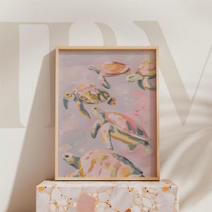 May include: A framed art print showing a watercolour painting of several sea turtles in shades of yellow, orange, and green. The turtles are set against a soft pink and blue background, creating a serene and artistic aesthetic. The frame is a light wood colour.