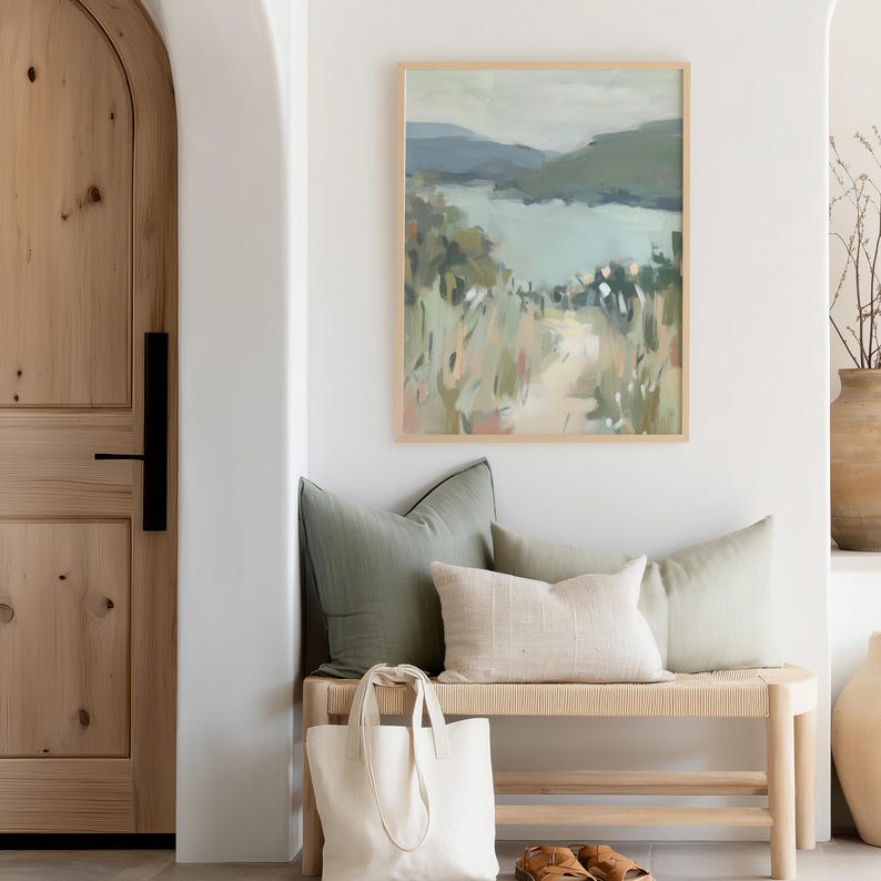 May include: A framed landscape painting with a light wood frame hangs above a bench with pillows. The painting depicts a lake and mountains in muted green and blue tones. A white tote bag sits on the bench.
