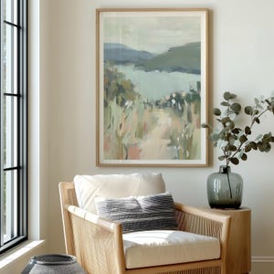 May include: A framed landscape painting with a light wood frame. The artwork depicts a path leading to a body of water, with green and blue hues dominating the scene. The painting hangs above a wicker chair with a white cushion and a decorative pillow.