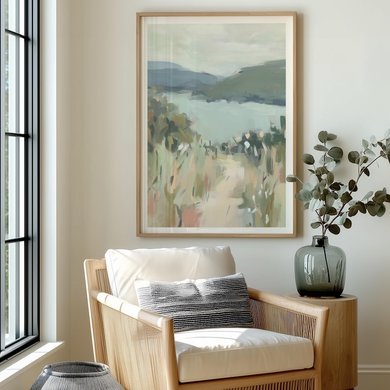 May include: A framed landscape painting with a light wood frame. The artwork depicts a path leading to a body of water, with green and blue hues dominating the scene. The painting hangs above a wicker chair with a white cushion and a decorative pillow.