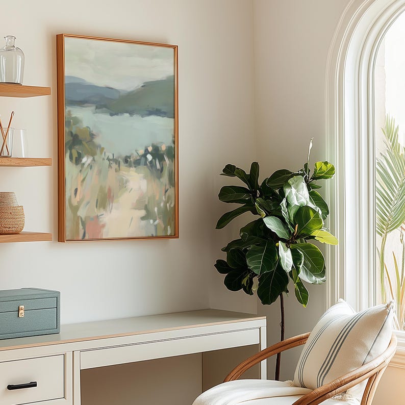 May include: An abstract landscape painting in a wooden frame hangs on a white wall. The artwork features muted tones of blue, green, and beige. A desk, a plant, and a chair are in the room. A decorative box and shelves are also visible.