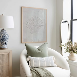 K&ouml;nnte beinhalten: Gerahmtes Wandbild mit einem wei&szlig;en Muscheldesign. Das Kunstwerk h&auml;ngt &uuml;ber einem wei&szlig;en Sessel mit gr&uuml;nen und gestreiften Kissen und einer gr&uuml;nen Decke. Eine blau-wei&szlig;e Lampe und ein Spiegel sind ebenfalls zu sehen.