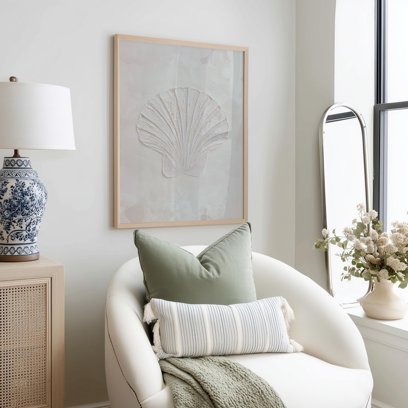 K&ouml;nnte beinhalten: Gerahmtes Wandbild mit einem wei&szlig;en Muscheldesign. Das Kunstwerk h&auml;ngt &uuml;ber einem wei&szlig;en Sessel mit gr&uuml;nen und gestreiften Kissen und einer gr&uuml;nen Decke. Eine blau-wei&szlig;e Lampe und ein Spiegel sind ebenfalls zu sehen.